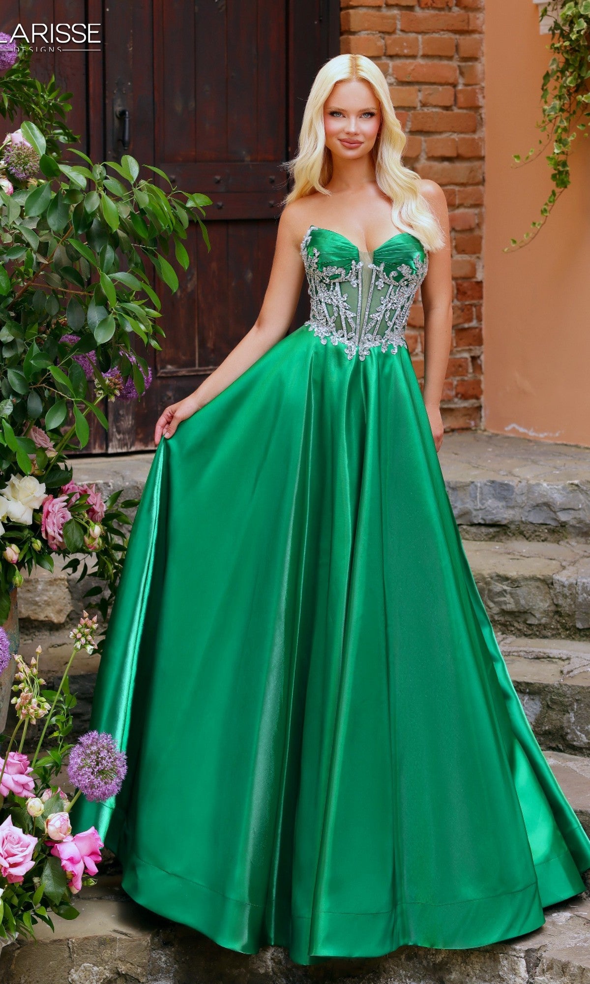 A woman in the Clarisse Designs 62140 Prom Ball Gown in emerald stands on stone steps near a brick wall and flowers, wearing a strapless dress with a beaded corset bodice.